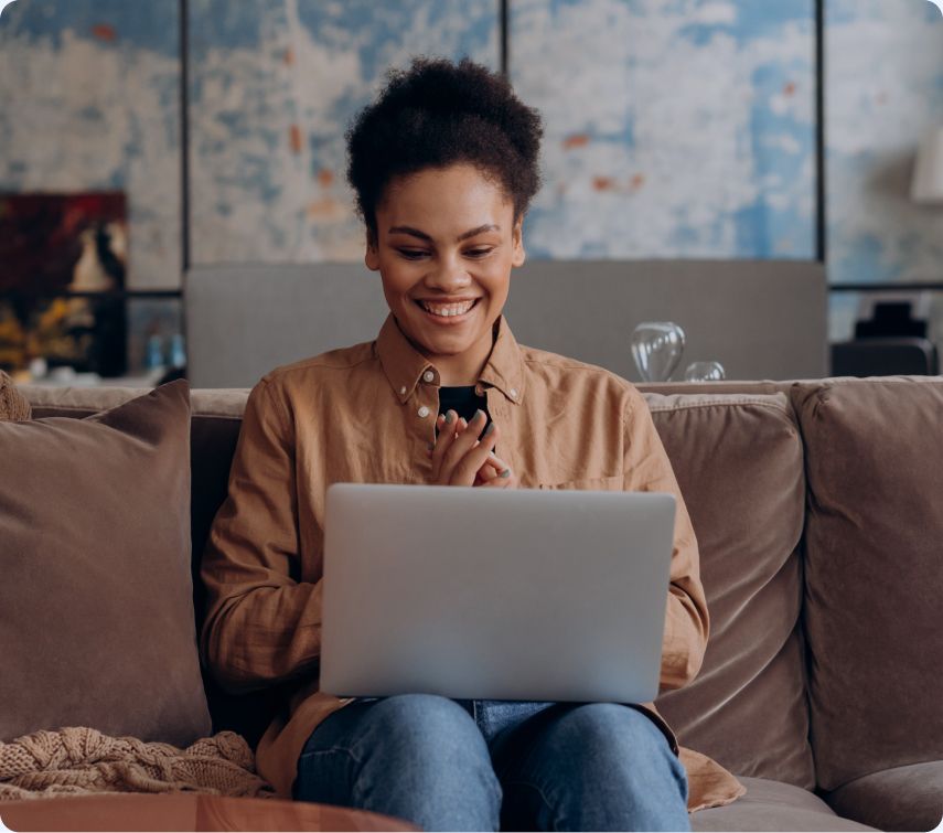 Photo of a person seated on a sofa with a laptop on their lap, smiling while looking at the screen in a living room setting.