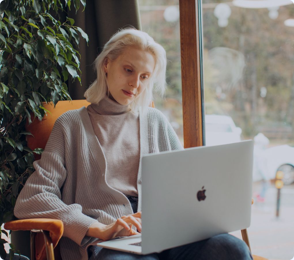 Photo of a person with light hair seated in a chair near a window, working on a laptop with a plant beside them.