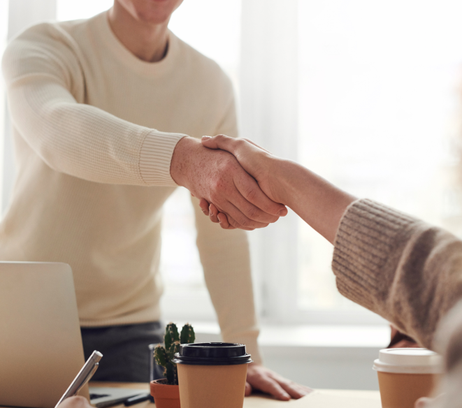 Handshake between two people at a desk with a laptop and coffee cups, representing a completed deal or partnership.