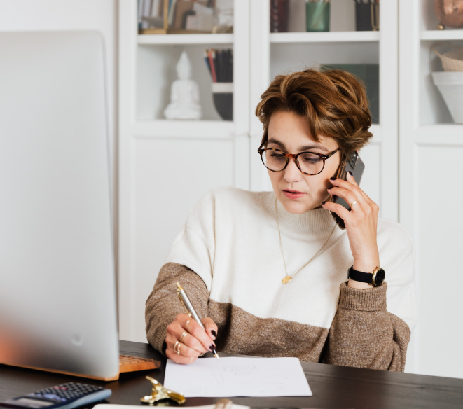 Person working at a desk, speaking on the phone and writing notes beside a laptop, representing customer support or professional consultation.